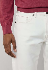 Jean en denim blanc avec des surpiqûres contrastantes, une poche avant avec des accents rouges et une coupe ajustée. La main repose légèrement sur le tissu.