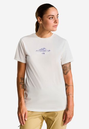 Frau mit Tattoos trägt ein weißes T-Shirt mit lila Bergmotiv und beige Hose und schaut nach rechts vor weißem Hintergrund.