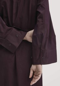 Person in einem dunkelviolettem langärmligen Kleidungsstück, mit der rechten Hand hinter dem Rücken und dem linken Arm entspannt nach unten an einem schlichten hellen Hintergrund.