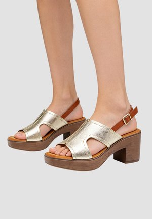 Pieds portant des sandales métalliques dorées à bout ouvert avec des brides marron à la cheville et des talons carrés en bois massif sur un fond neutre.