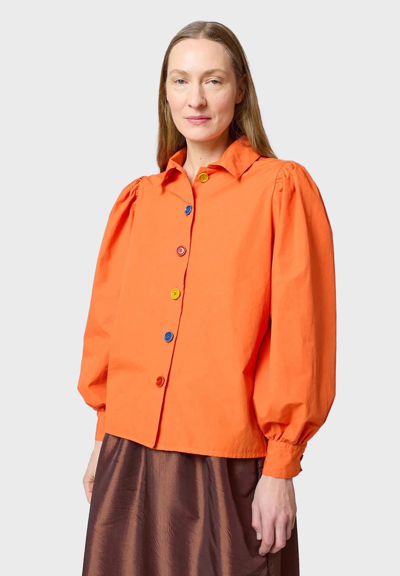 Kirkkaan oranssi nappipaita puffihihnoilla, jossa on moniväriset napit. Suuri puuvillakangas, lyhennetty malli, yhdistetty ruskeisiin hameisiin.