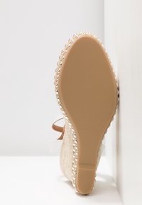 Semelle d'une sandale compensée beige avec des côtés en corde tressée et des lanières blanches, photographiée debout contre un mur d'angle blanc.