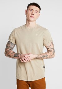 Beige katoenen t-shirt met korte gerolde mouwen, ronde halslijn en een klein logo op de borst. Heeft zichtbare tatoeages op de armen.