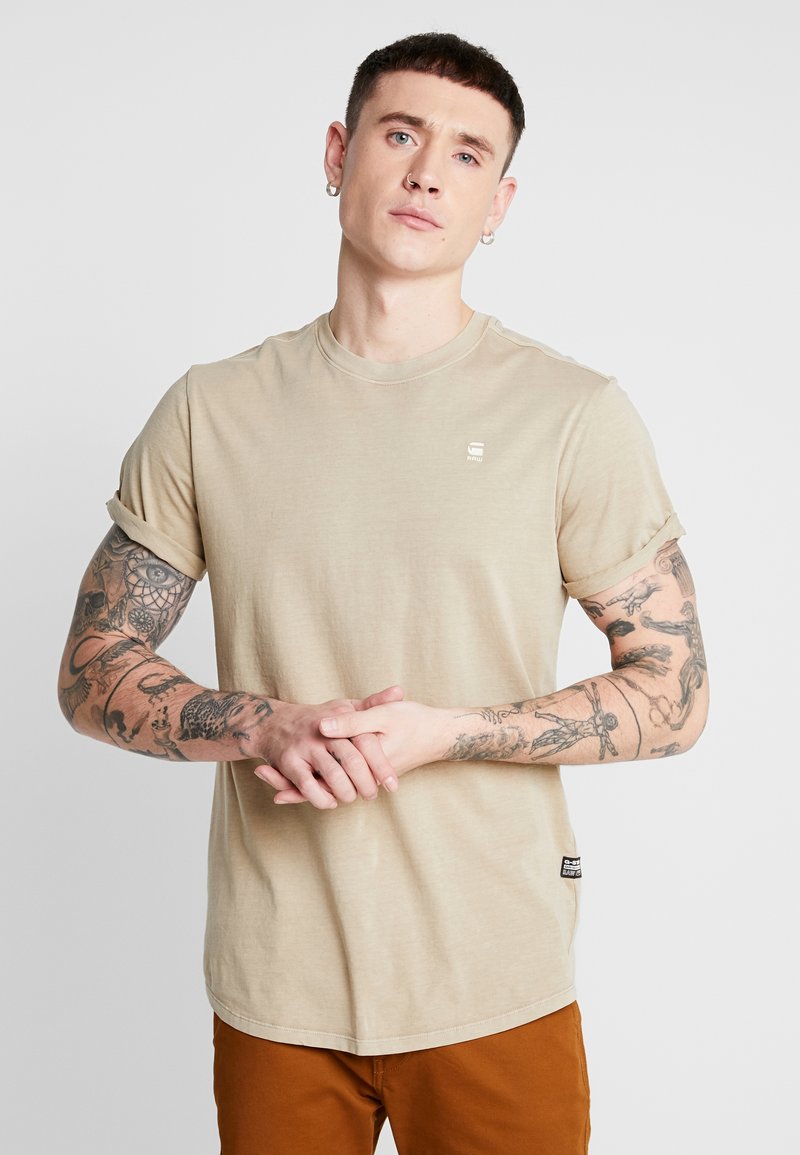 Beige katoenen t-shirt met korte gerolde mouwen, ronde halslijn en een klein logo op de borst. Heeft zichtbare tatoeages op de armen.