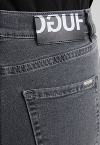 Jeans de mezclilla gris con una superficie texturizada, una etiqueta de logo prominente en la cinturilla y un pequeño acento de hardware plateado en el bolsillo trasero.