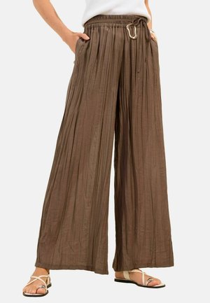 Pantaloni maro cu talie largă plisată, prevăzuți cu șnur și cataramă metalică, purtați cu sandale albe și un top alb.