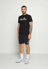 T-shirt preta com logótipo "ellesse" cinzento e detalhe a cores, combinado com calções cinzentos com logótipo e riscas brancas, usado com ténis.