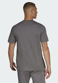 Graues Baumwoll-T-Shirt mit kurzen Ärmeln und Rundhalsausschnitt. Glatte Textur und lockere Passform, getragen von einer Person von hinten mit sichtbarem Tattoo.