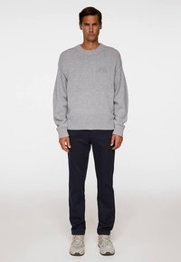 Jasnoszary sweter z dzianiny z prążkowanym dołem i mankietami, zestawiony z ciemnoniebieskimi spodniami. Model nosi beżowe trampki. Prosty design, bez wzorów.