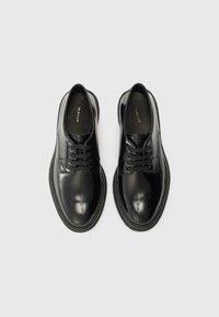 Chaussures en cuir noir avec une finition brillante, bouts ronds, design à lacets et semelles en caoutchouc ; marque visible à l'intérieur de la chaussure.