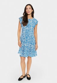 Robe bleue avec un imprimé léopard abstrait blanc, manches courtes à volants, taille élastique et jupe évasée. Portée avec des chaussures plates noires.