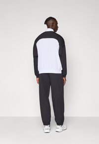 Sorte og hvide atletiske løbebluser med elastiske manchetter og krave, parret med sorte joggers. Bemærkelsesværdig logo detalje på ryggen.