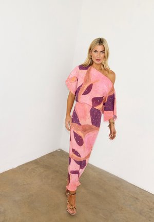 Ξανθιά γυναίκα που στέκεται στη γωνία φορώντας ροζ και μοβ ριγέ φόρεμα με έναν ώμο και πλατφόρμες με λεοπάρ τύπωμα σε δάπεδο από σκυρόδεμα.