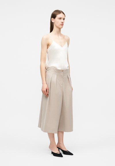 Žena oblečená v béžových širokých nohaviciach nad členky, bielej hodvábnej tielkovine a čiernych topánkach s nízkym podpätkom a špicatou špičkou, stojaca na jednoduchom pozadí.