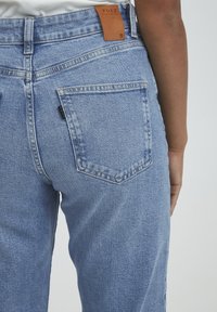 Jeans de mezclilla azul claro con un bolsillo trasero y una etiqueta de marca de cuero marrón en la cintura. La tela tiene un acabado texturizado.