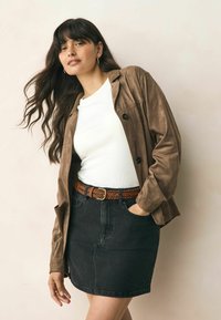 Brun mockaläderjacka över en vit ribbad skjorta, tillsammans med en svart denimkjol och ett flätat brunt läderbälte. Mjuk textur genom hela outfiten.