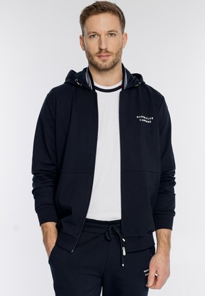 Mężczyzna ubrany w czarną bluzę z kapturem zapinaną na zamek z napisem "Marine Club Campione", białą koszulkę oraz czarne spodnie ze sznurkiem, stojący ze schowanymi w kieszeniach rękami.