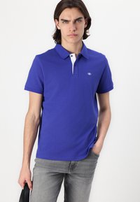 Blå polo skjorte med hvid krave, korte ærmer og et lille logo på brystet. Bæret sammen med lysegrå jeans.