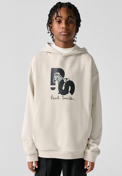 Jeune personne aux cheveux tressés portant un sweat à capuche beige Paul Smith avec un motif zèbre noir et un pantalon noir, debout devant un fond uni.
