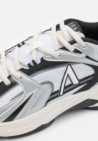 Sneaker bianco in rete con dettagli in pelle nera e grigia. Presenta lacci beige, dettaglio del logo argentato e suola testurizzata.
