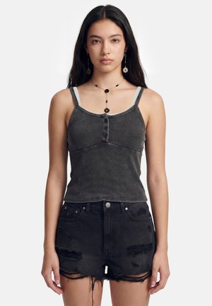 Ung kvinde med langt mørkt hår iført sort ærmeløs top med knapper, sorte slidte denimshorts og blomsterøreringe og halskæde.