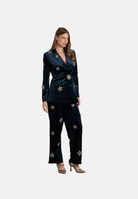 Marineblauwe fluwelen pak met een op maat gemaakt blazer en losse broeken, versierd met gouden stervormige applicaties en gecombineerd met metallic hakken.