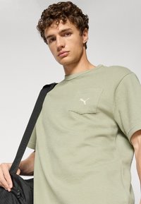 Lichtgroen t-shirt met korte mouwen en een textuurpatroon, voorzien van een linkerkant borstzak en een klein wit logo. Het model houdt een zwarte tas vast.