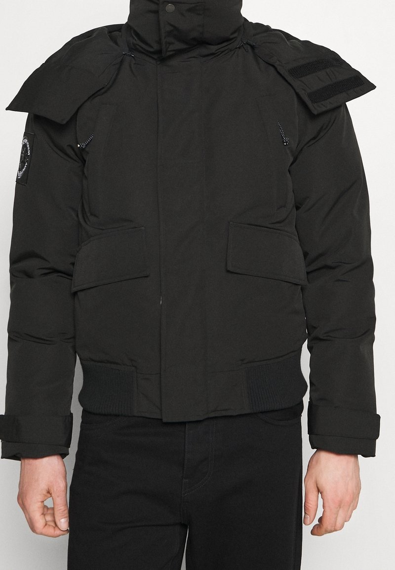 Sort polstret jakke med høj krave, skulderdetaljer med klap, forlommer med klapper, ribbet taljebånd, båret med sorte bukser.