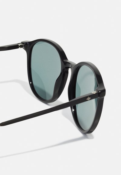 Mustat pyöreät aurinkolasit vihreillä linsseillä. Muovikehyksissä on tyylikäs muotoilu ja tekstuuriset templet. Huomiota herättävänä yksityiskohtana on hopeinen yksityiskohta.