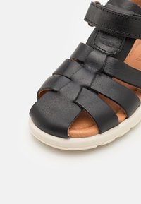 Musta nahkainen sandaali, jossa on punottu remmimalli, avoin varvas, beige ulkopohja ja ruskea sisävuori. Säädettävä hihna varmistaa tukevan istuvuuden.