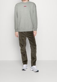 Persona che indossa una felpa grigia Levi's, pantaloni in corduroy marroni e sneakers bianche, in piedi contro uno sfondo bianco semplice.