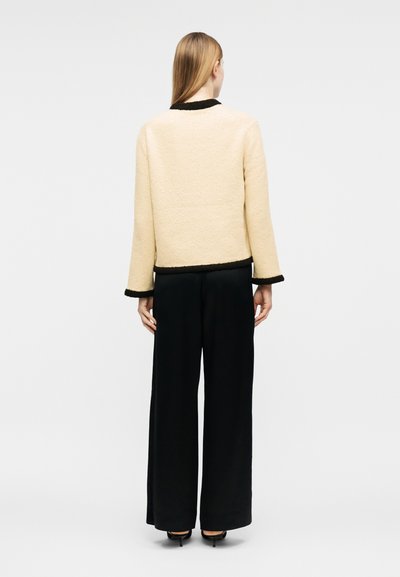 Femme aux cheveux blonds raides portant une veste beige texturée avec des bordures noires et un pantalon noir à jambes larges, debout de dos.