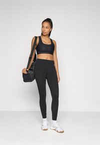 Kvinne i svart sport-bh og tights som bærer treningsbaggen over skulderen, stående mot en enkel lys bakgrunn.