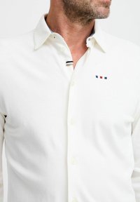 Cămașă albă cu mânecă lungă, confecționată dintr-un material moale, cu guler căptușit. Prezintă un mic logo brodat în albastru, roșu și bleumarin.