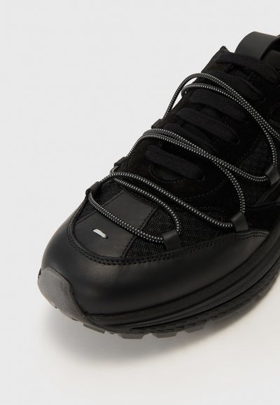 Black sneaker with a smooth leather toe cap, textured mesh upper, reflective laces, and a cushioned sole. Notable grey accents on the heel.