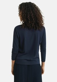 Navy blue pullover sweater with long sleeves and ribbed cuffs, paired with a pleated dark skirt, shown from the back. Smooth texture.