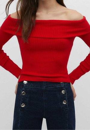 Femme portant un haut rouge côtelé, à manches longues et épaules dénudées, avec un jean bleu foncé taille haute à grands boutons argentés sur les côtés.