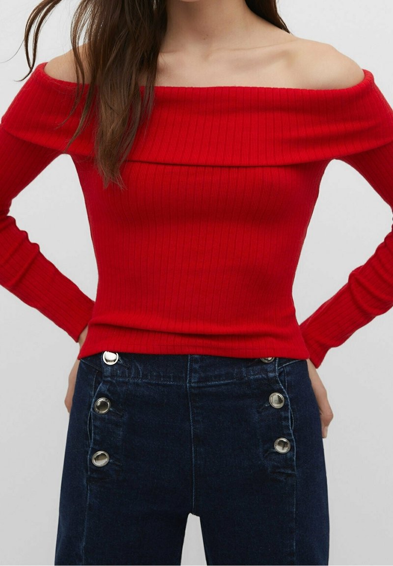 Femme portant un haut rouge côtelé, à manches longues et épaules dénudées, avec un jean bleu foncé taille haute à grands boutons argentés sur les côtés.