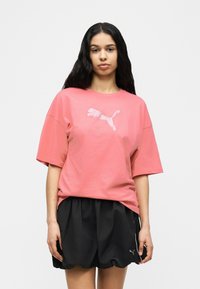 Γυναίκα με μακριά μαύρα μαλλιά, που φοράει ροζ μπλούζα Puma και μαύρο σορτς Puma, στέκεται μπροστά σε απλό ανοιχτό φόντο.