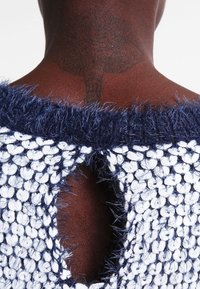 Marinblå och vit stickad plagg med en texturerad, pälsliknande krage, med en urklippt detalj på ryggen mot en slät hudbakgrund.