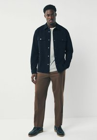 Camisa azul marino de botones con dos bolsillos en el pecho sobre una camiseta de rayas blanca y verde, combinada con pantalones marrones y zapatos negros de cordones.