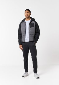 Gevoerde jas met een grijze voorkant, zwarte mouwen en een ritszak op de borst. Combinatie met zwarte broek en lichtgrijze sportschoenen.