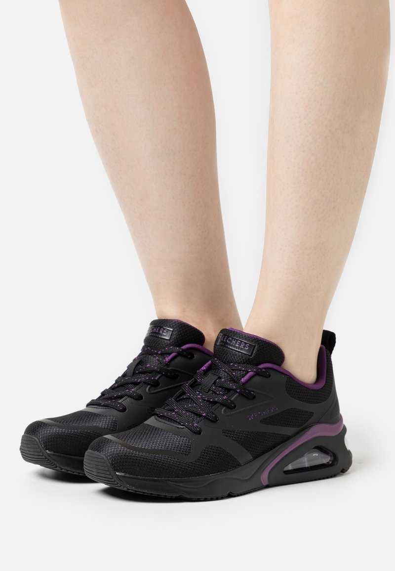Baskets de sport noires et violettes avec tige en mesh et semelles en caoutchouc, portées pieds nus, montrant les angles latéraux et avant.