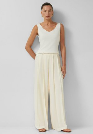 Kobieta w białym topie bez rękawów z dekoltem w serek, szerokich kremowych spodniach, złotych kolczykach kołach i brązowych sandałach, stojąca przy szarej ścianie.