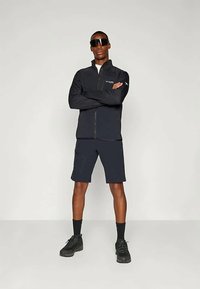 Μαύρο φούτερ με φερμουάρ και ανακλαστικές λεπτομέρειες, συνδυασμένο με σκούρες βερμούδες και μαύρα αθλητικά παπούτσια. Το μοντέλο στέκεται με αυτοπεποίθηση με τα χέρια σταυρωμένα.