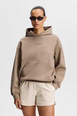 Ženska, oblečena v bež črtasto jopico s sprednjim žepom in krem kratke hlače, dopolnjena z ozkimi črnimi sončnimi očali, stoji ob navadni ozadju.