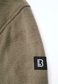 Olive roheline fliisjakki kangas, millel on pehme, tekstuuriga pind. Varrukal on must silt valge "B" logoga.