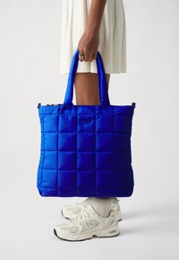 Sac de toile matelassé bleu avec texture rembourrée, poignées de transport doubles et fermeture zippée. Associé à des baskets de couleur claire.