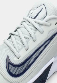 Close-up of white athletic shoe with textured fabric, navy blue swoosh logo, and white laces tied over padded tongue.