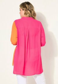 Camisa larga en rosa brillante y naranja, hecha de una tela ligera. Presenta un cuello, un plisado en la parte posterior y se combina con pantalones blancos.